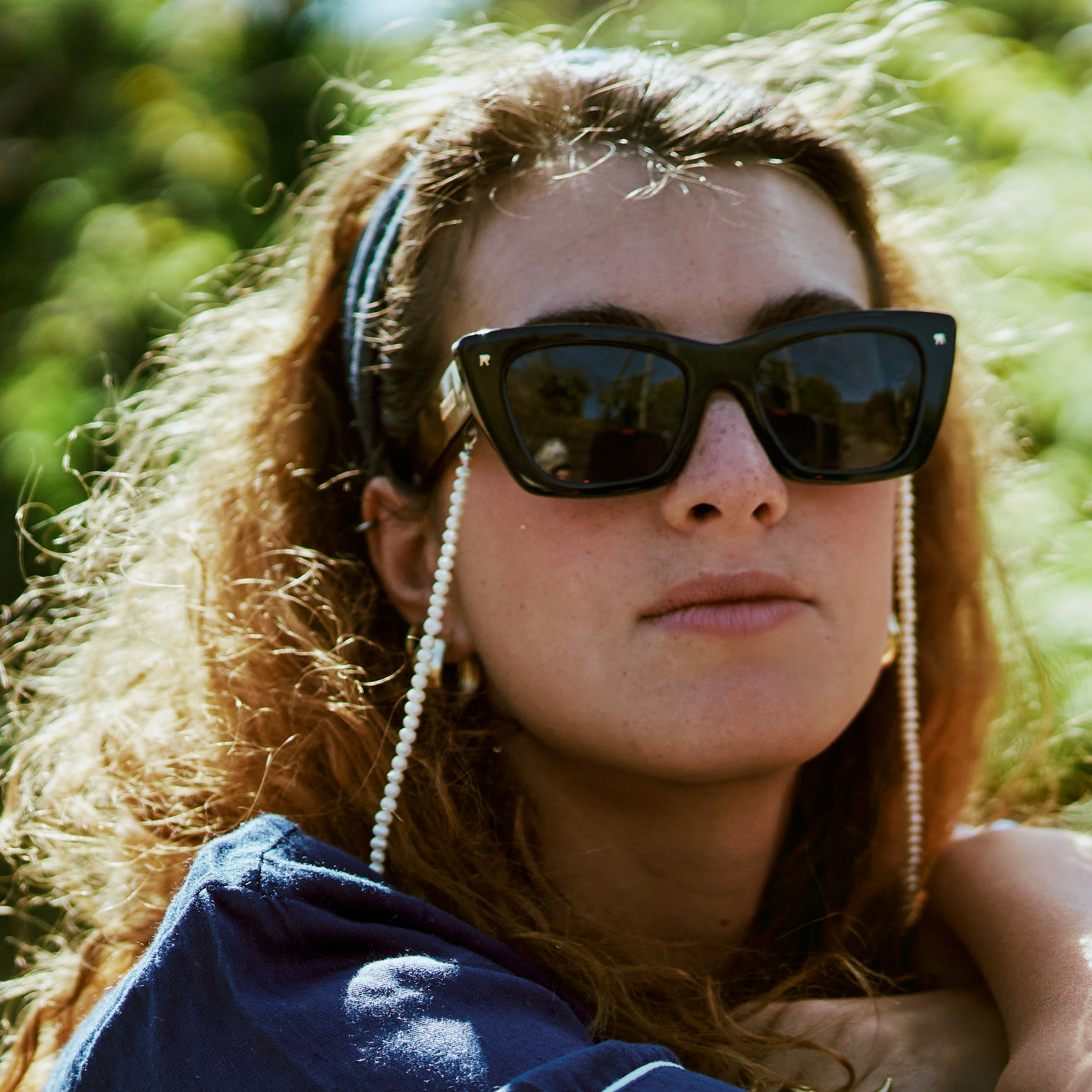 Woman wearing sunglasses with chain, blue headband, and blue top outdoors.